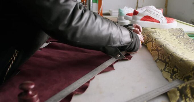 Footwear manufacturing. Shoes factory. Unrecognizable craftsman hands using stationery knife metal ruler cutting material at workspace.