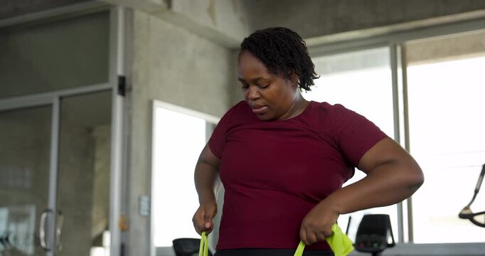 African American plus size woman pulls resistance band near waist with elbows bent during strength exercise in modern gym. Curvy female athlete trains arms, shoulder, endurance, confidence, wellness.