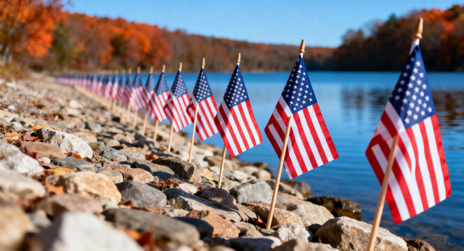 A row of American flags placed along a rocky lake shore with vibrant autumn trees in the background during a peaceful sunny day in fall