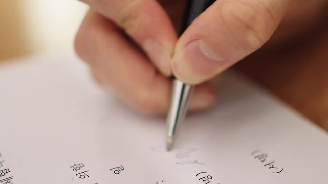 A close-up of a hand holding a pen and writing cursive text or a signature on white paper in a warm indoor setting.