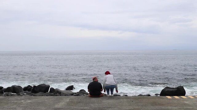 Woman walking along a scenic seaside promenade to a man, surrounded by rocks and the gentle waves of the ocean. The relaxed and peaceful atmosphere is evident as people enjoy.