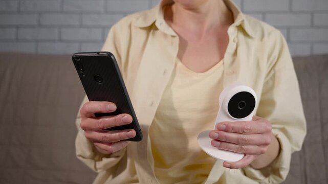 Woman connecting smartphone to smart home security camera. Unrecognizable woman holding a modern smartphone and a wireless indoor surveillance camera, setting up a home security