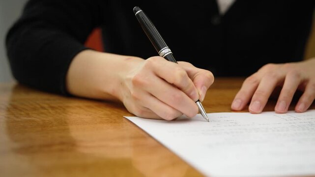 A close-up of a woman's hand signing a document with a pen on a wooden desk surface.