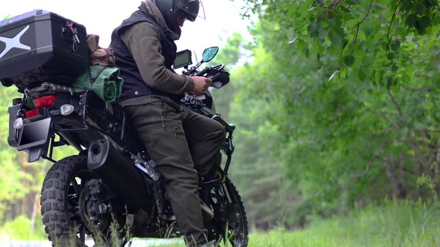Middle-aged motorcyclist stands on the side of a country road, leaning against his touring motorcycle, scrolling through his smartphone's news feed and using GPS. Relaxing after a long journey.