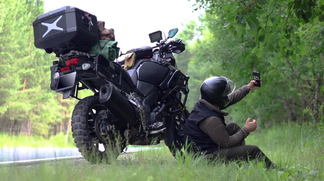 Motorcyclist sitting on the side of a country road, next his touring motorcycle and taking a selfie. Relaxing after a long journey. Slow motion 4K footage.