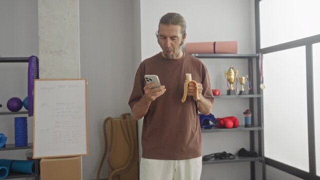 Man holding smartphone and banana, looking at phone in fitness studio with yoga mats, ropes, boxing gloves and trophies on shelf; calm routine.