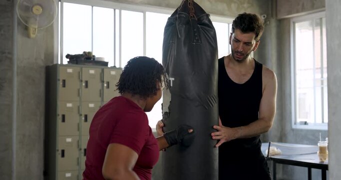 Plus size African American woman learns boxing with male trainer in modern gym, facing punching bag during partner coaching session, improving punch technique, cardio endurance, strength, motivation.
