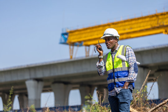 Construction engineer with blueprints using a walkie talkie at a highway infrastructure large scale project, Site manager wearing hard hat and safety vest communicating at a bridge construction site