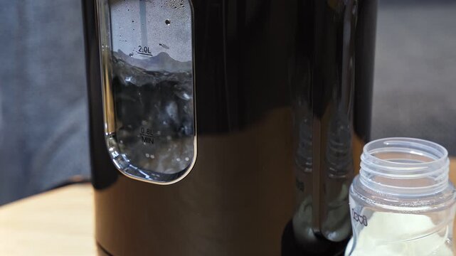 a black hot water dispenser boiling water next to a clear plastic baby bottle containing white formula powder. The domestic indoor scene highlights everyday modern parenting routines.