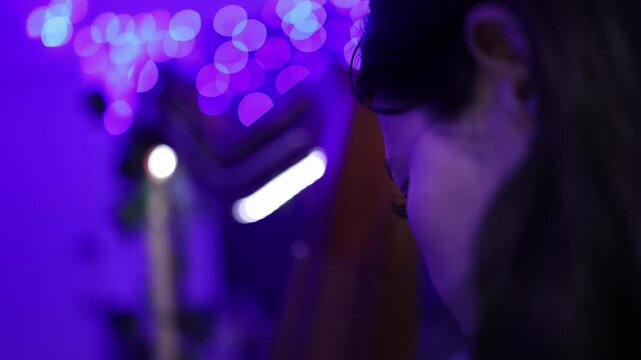 Artistic close-up shot of a woman playing a harp beneath vibrant purple bokeh lights. Soft focus, moody ambiance, and cinematic lighting create a serene, intimate music moment.