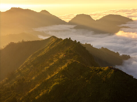 Aerial view of Gunung Bismo at sunrise with golden light illuminating the mountain ridges and a sea of clouds in the valley in Kecamatan Kejajar, Jawa Tengah, Indonesia.