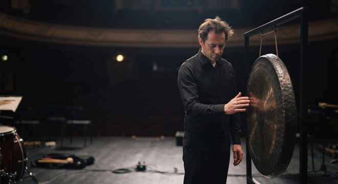 A candid, contemplative photograph of a percussionist standing alone on a silent, darkened stage after a powerful performance, resting a hand on the edge of the still-vibrating gong,