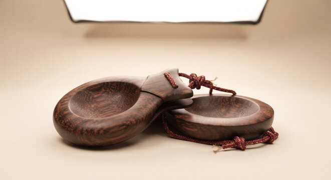 A professional product studio shot of a pair of traditional hand-held castanets, presented against a seamless, minimalist cream-colored background