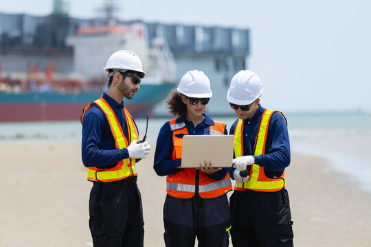 Team of diverse engineers in hard hats and safety vests collaborates on beach, using laptop and radio with large cargo ship in background