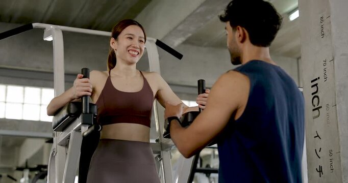 Young Asian couple preparing gym exercise together, woman holds handles on knee raise station while man explains technique, partner training with support, coaching, motivation, wellness lifestyle.