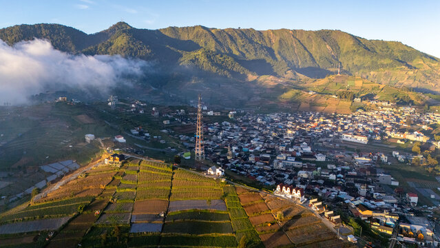 Aerial view of the Dieng Plateau featuring terraced agricultural fields, a small town nestled in the valley, a communication tower, and rolling mountains in Indonesia.