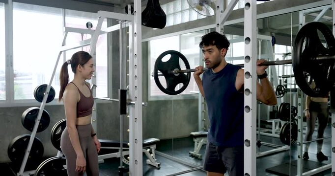 Young Asian couple practicing barbell squat in modern gym, man demonstrates rack movement while woman watches nearby, partner workout coaching with support, motivation, leg strength, wellness routine.