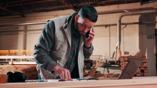 Carpenter, laptop and phone call in workshop for timber distribution, communication or stock deadline. Male person, artisan or talking with smartphone or computer for lumber production in warehouse