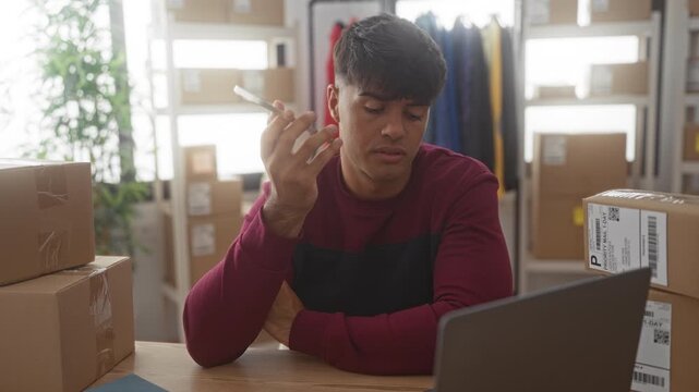 Man holding smartphone to ear among stacked shipping boxes and laptop in a warehouse building, checking orders on a laptop; concern responsibility.