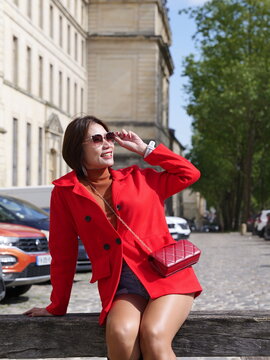 A woman wearing a vibrant red coat and sunglasses sits on a wooden beam along a city street with parked cars and classic architecture in the background