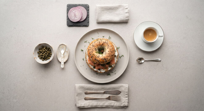 A symmetrical top-down "flat lay" of a classic NYC bagel setup. The lox bagel is the centerpiece, flanked by an organized array of a small bowl of capers, a few slices of red onion