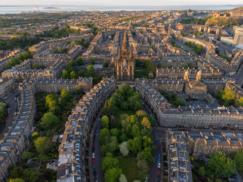 Aerial view of St Mary's Cathedral with its gothic spires surrounded by historic stone buildings, lush trees, and urban streets during the golden hour in Edinburgh, Scotland, United Kingdom.