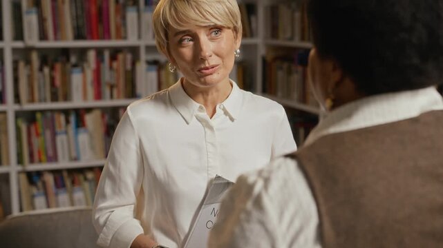 Middle-aged Caucasian woman holding book titled New Chapter and talking to African American colleague in library before hugging. Perfect for book publishing, career coaching, or education promos