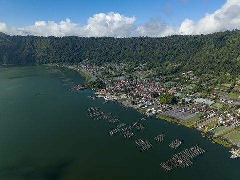 Aerial view of Lake Batur with its surrounding lush green mountains, traditional village houses along the shoreline, and floating fish farms in the water in Kintamani, Bali, Indonesia.