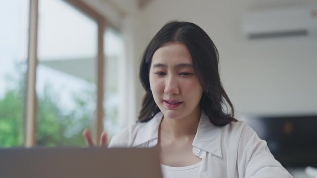 Asian woman using laptop for online video conference with her team, working remotely from home, talking and discussing work in a living room