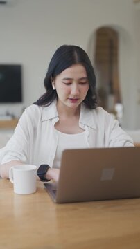 Young Asian woman using laptop with wireless earbuds for online meeting, working remotely from home at wooden table with coffee mug, talking with her team