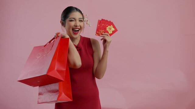 Excited young Asian woman is holding shopping bags and angpao or red packet monetary gift, isolated by pink background.
