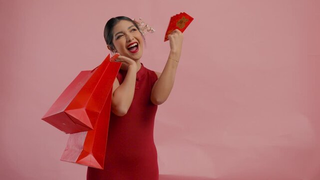 Excited young Asian woman is holding shopping bags and angpao or red packet monetary gift, isolated by pink background.