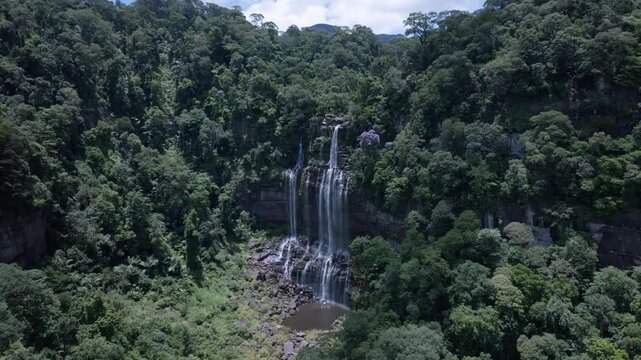 A drone flying over a waterfall in a deep green forest provides an aerial, top-down view of a National Park in Laos PDR.