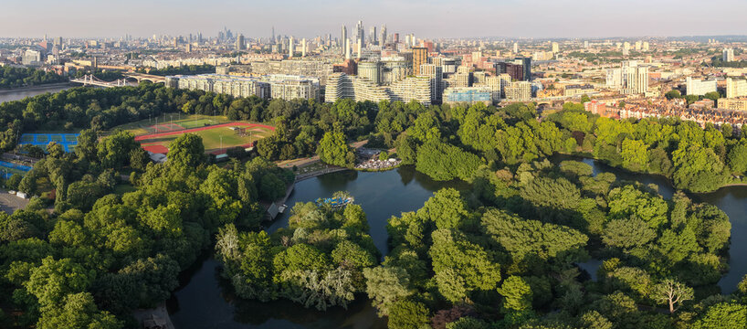An ultra-wide panoramic aerial view looking across the lush green treetops, boating lake, and recreational sports grounds of Battersea Park toward the distant London cityscape.