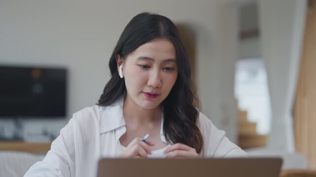 Asian woman wearing wireless earbuds speaking with an AI voice assistant on laptop, using chatbot technology to work from home, talking and listening in a bright room