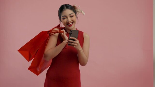 Smiling young Asian woman in a traditional cheongsam dress is holding smartphone and shopping bags, isolated by pink background.