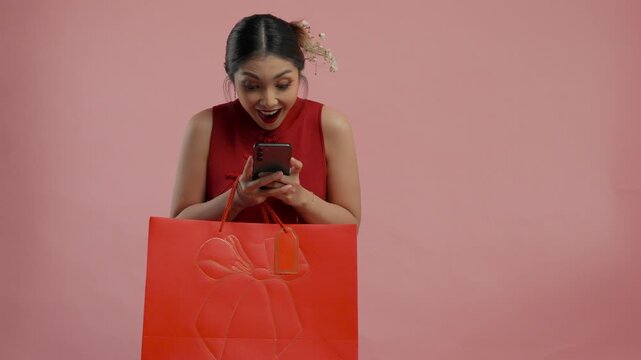 Shocked young Asian woman in a traditional cheongsam dress is holding smartphone and shopping bags, isolated by pink background.