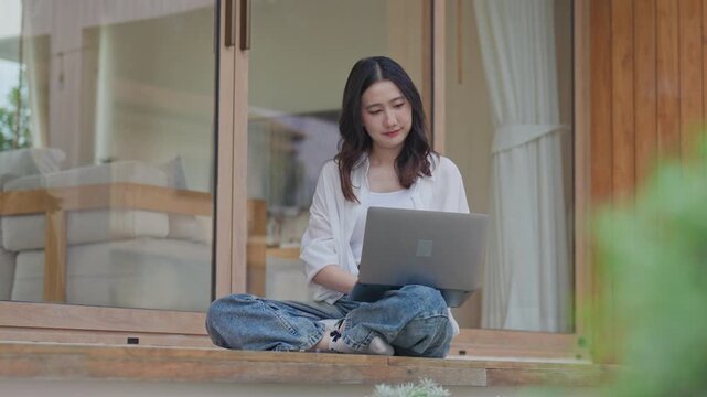 Asian woman sitting cross legged on wooden terrace working on laptop at modern minimal home, enjoying flexible remote work and a calm slow living lifestyle outdoors