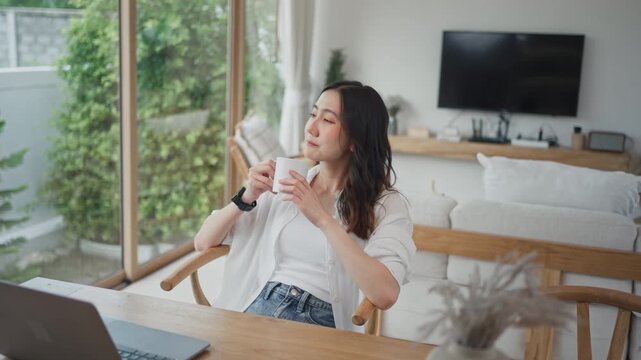 Asian woman taking a coffee break while working from home on laptop, relaxing in a chair at modern minimal living room, enjoying a calm moment between work tasks