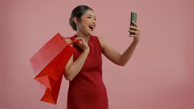 Happy young Asian woman in a traditional cheongsam dress looking at her smartphone while shopping, isolated by pink background.