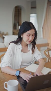 Asian woman wearing earbuds typing on laptop and writing notes in notebook, working from home and planning tasks, organizing ideas in a bright modern room