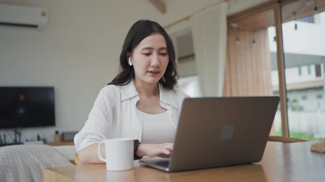 Asian woman wearing earbuds typing on laptop and writing notes in notebook, working from home and planning tasks, organizing ideas in a bright modern room