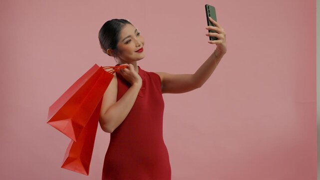 Happy young Asian woman in a traditional cheongsam dress looking at her smartphone while shopping, isolated by pink background.