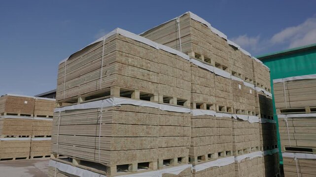 Large bundles of cut wooden boards are stacked on pallets outdoors under a blue sky, ready for storage, shipping, or construction supply