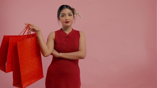 Pretty young Asian woman in traditional cheongsam dress is smiling and giving shopping bags, isolated by pink background.