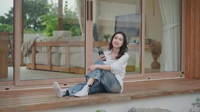 Asian woman sitting on wooden terrace of modern minimal home holding smartphone, relaxing on a calm weekend, enjoying a slow living moment in a cozy living space
