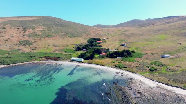 Aerial drone footage of a small remote settlement beside a sheltered turquoise bay and rolling treeless hills in the Falkland Islands. Colors: turquoise, green, beige.