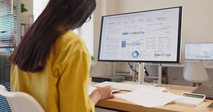 Woman analyst accountant reviews finance analytics report. At a computer monitor in an office, she reviews a spreadsheet and printed pages for budgeting. Financial data guides smart choices.
