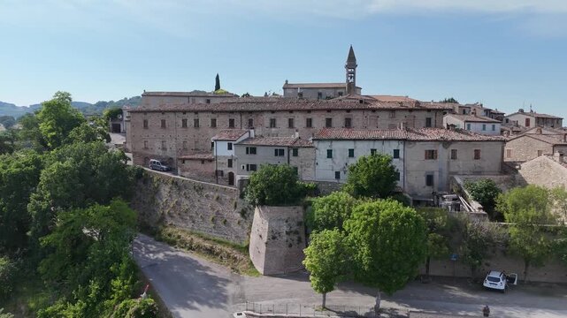 Borgo collinare con case in pietra e tetti in terracotta, dominato da una torre campanaria e circondato da alberi e campagna verde sotto cielo azzurro