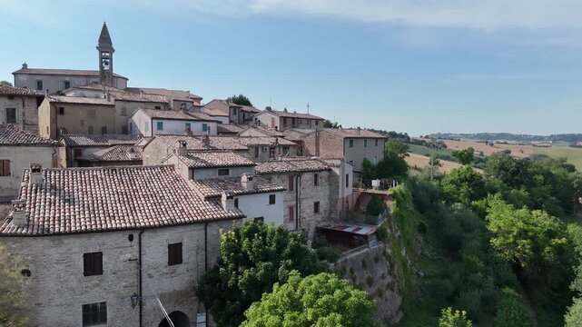 Borgo collinare con case in pietra e tetti in terracotta, dominato da una torre campanaria e circondato da alberi e campagna verde sotto cielo azzurro